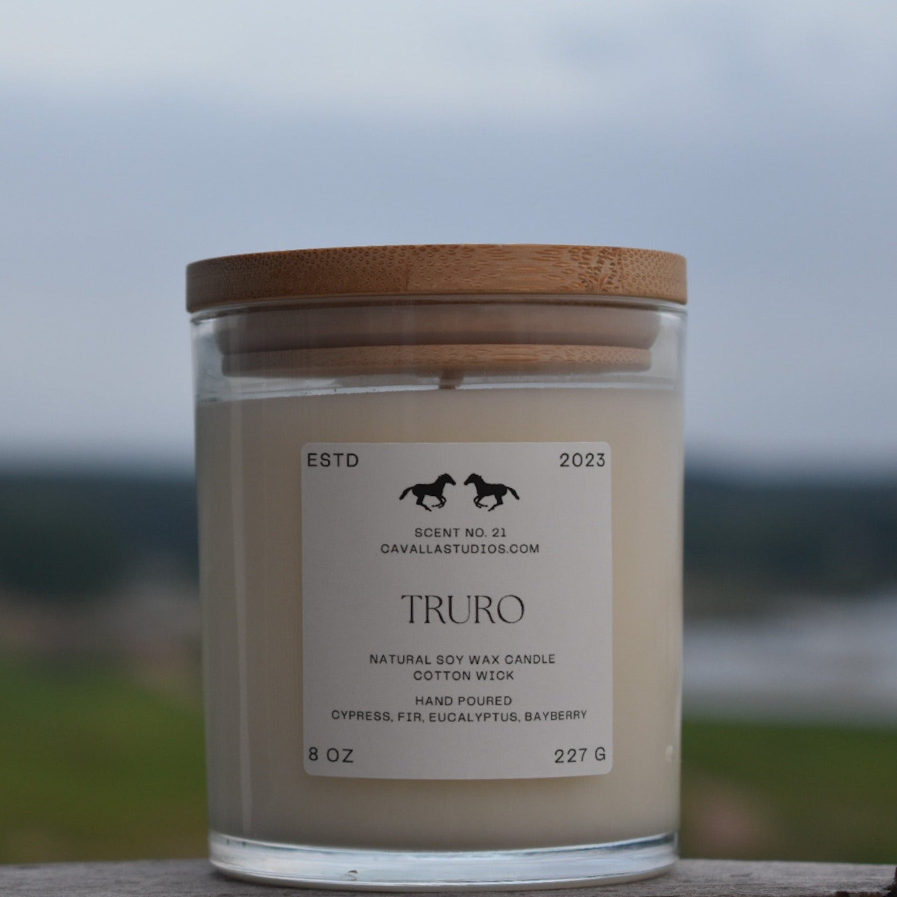 Classic white candle in a clear glass vessel covered by a bamboo lid. The glass features the 'Cavalla Studios' logo and the candle is labeled with the scent name 'Truro.' The blurred background is Pamet River.