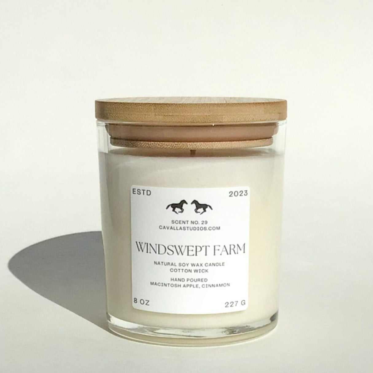Classic white candle in a clear glass vessel covered by a bamboo lid. The glass features the 'Cavalla Studios' logo and the candle is labeled with the scent name 'Windswept Farm.'
